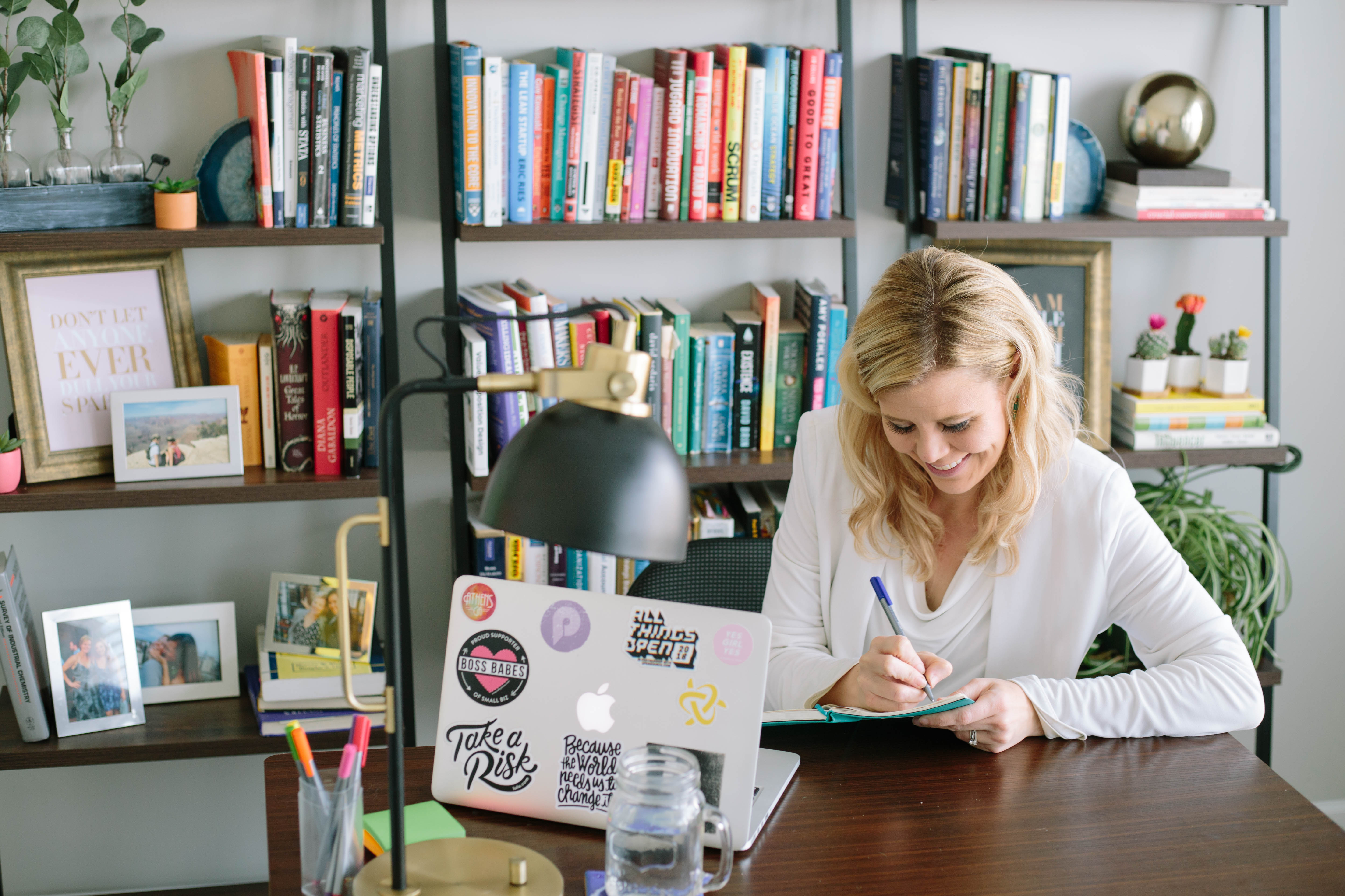 blond haired woman at desk taking notes with bookshelf behind her