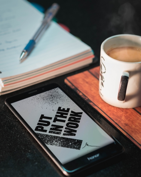 ipad on desk with image that says 'put in the work' along with a coffee mug and pad with pen